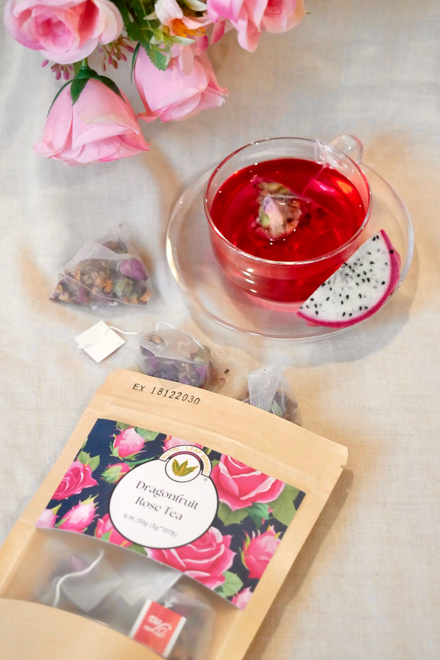Tea bag packaging with a dragon fruit tea label, a cup of red tea, and pink flowers on a light surface.